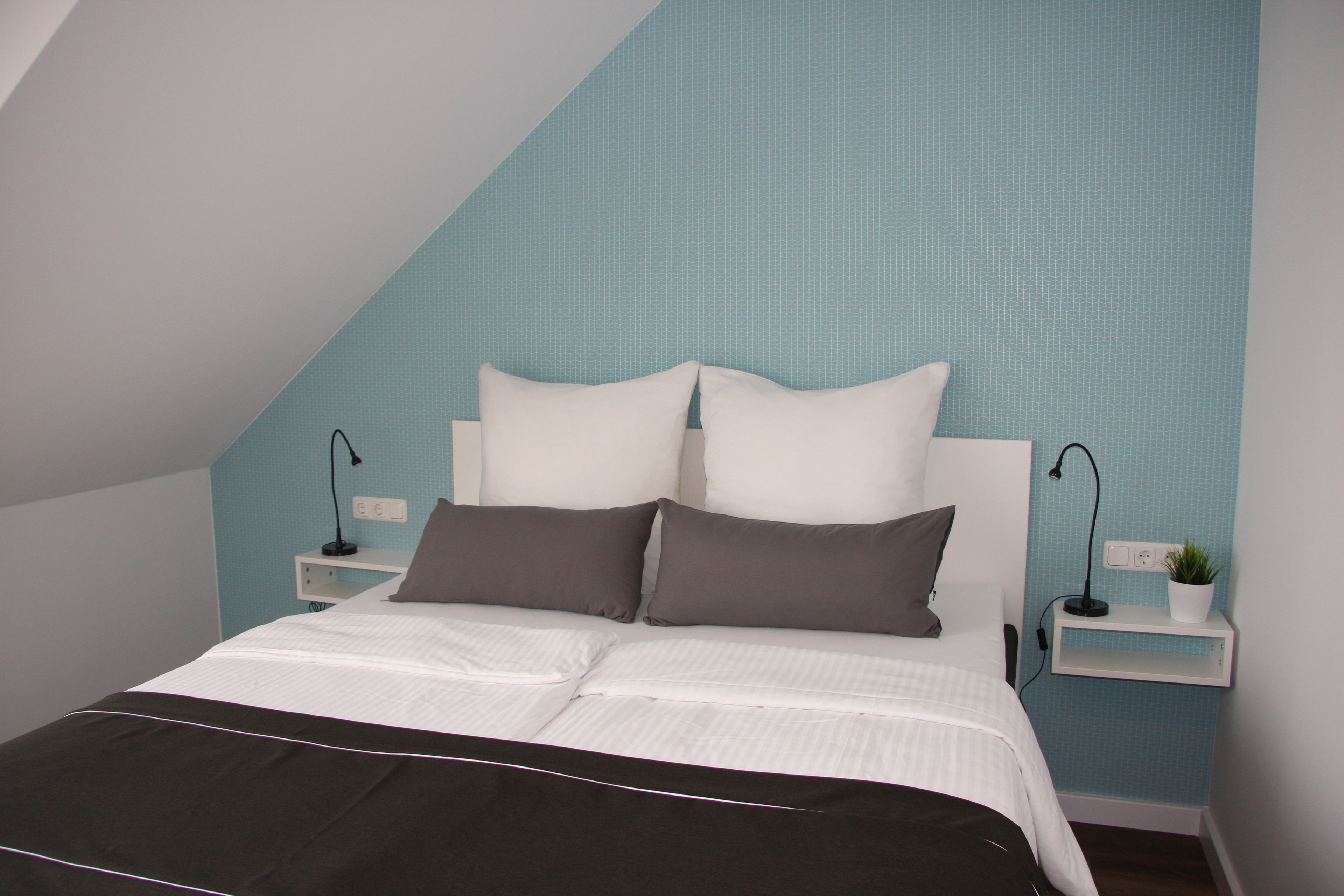 Ein Schlafzimmer mit einem Bett, einem weißen Kopfende, zwei Nachttischen mit Lampen, einer kleinen Pflanze und einer blauen, strukturierten Wand.