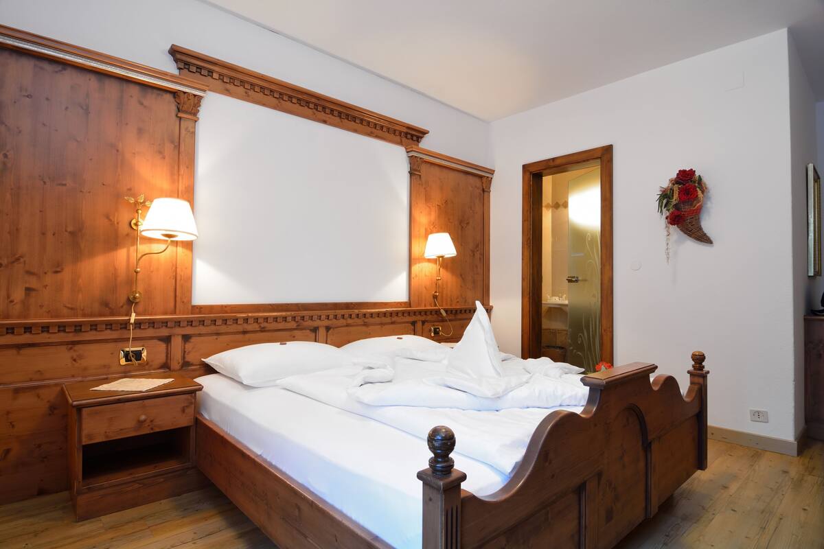 A wooden four-poster bed with white linens, two bedside lamps, a wooden nightstand, and a mirror on the wall.