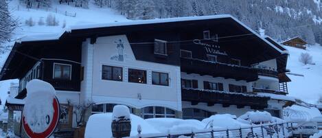 A hotel with a snow-covered exterior, a sign reading "Garni Wildbach," and a view of a snowy mountain in the background.
