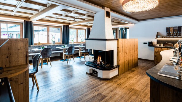 A modern interior with a central fireplace, wooden ceiling, and a dining area with natural light. - Garni Wildbach (Selva di Val Gardena)
