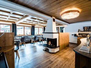 A modern interior with a central fireplace, wooden ceiling, and a dining area with natural light. - Garni Wildbach (Selva di Val Gardena)