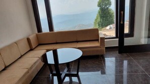 A room with a beige sofa, a small round table, and a large window offering a view of the outdoors. - Parwati Retreat (Ranikhet)