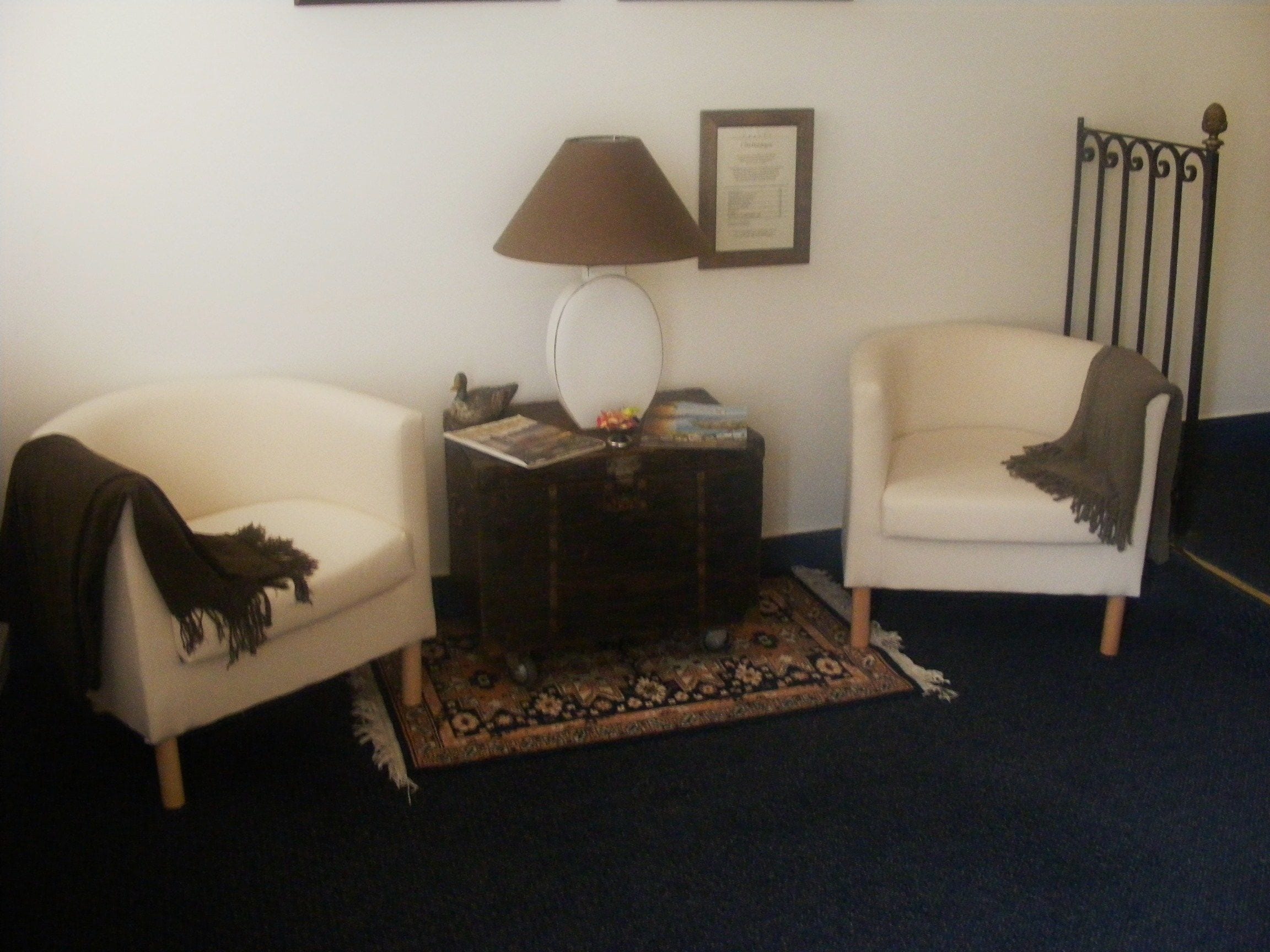 Un salon avec deux fauteuils blancs, une table d’appoint en bois foncé sur laquelle se trouve une lampe, un certificat encadré accroché au mur et un tapis à motifs.