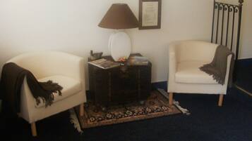 Un salon avec deux fauteuils blancs, une table d’appoint en bois foncé sur laquelle se trouve une lampe, un certificat encadré accroché au mur et un tapis à motifs.
