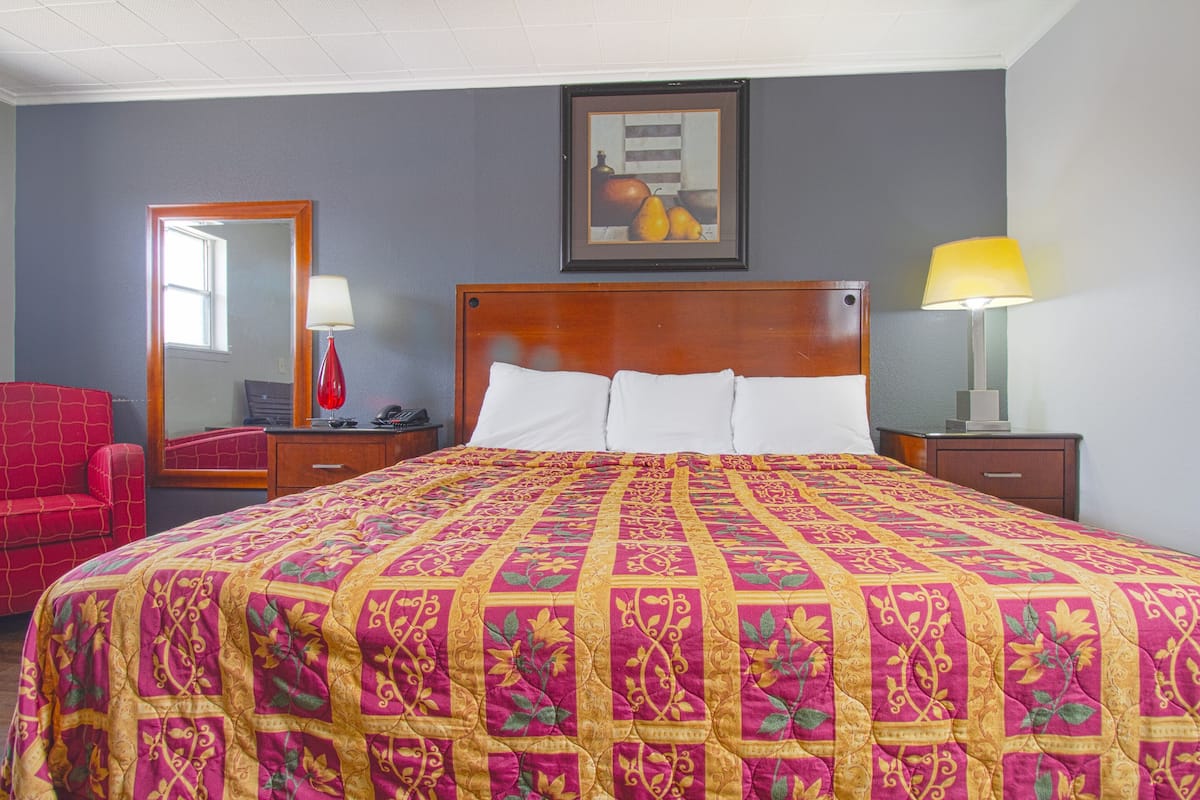 A hotel room with a large bed, a wooden headboard, a red armchair, a nightstand with a lamp, and a framed artwork on the wall.