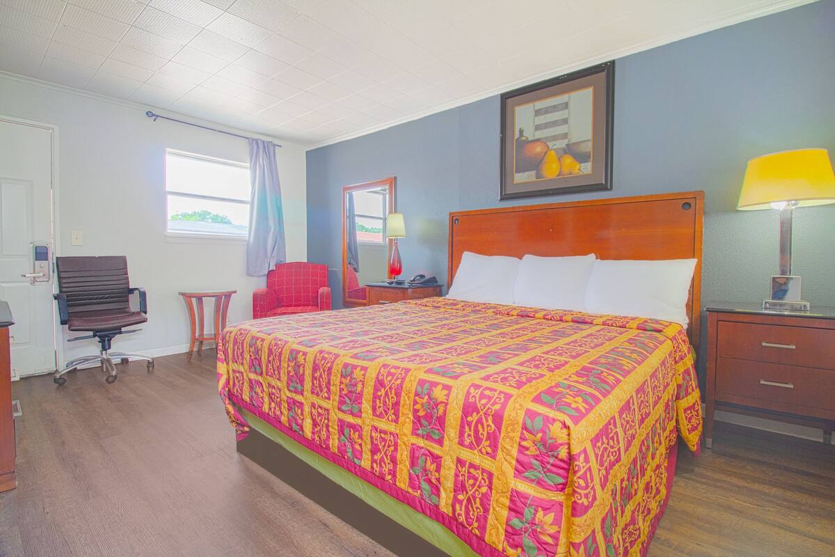 A hotel room with a large bed, a desk, a chair, a lamp, and a window.