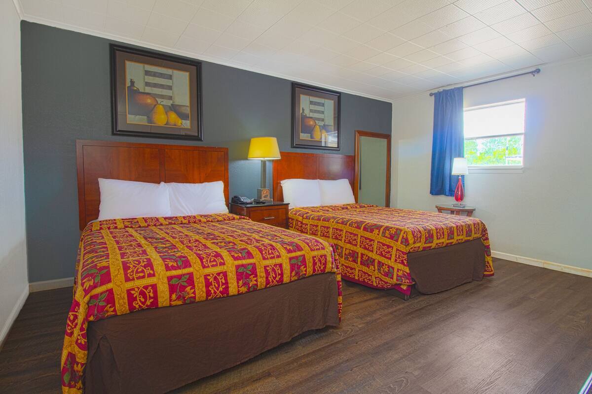 A hotel room with two beds, wooden headboards, a nightstand with a lamp, and a window with curtains.