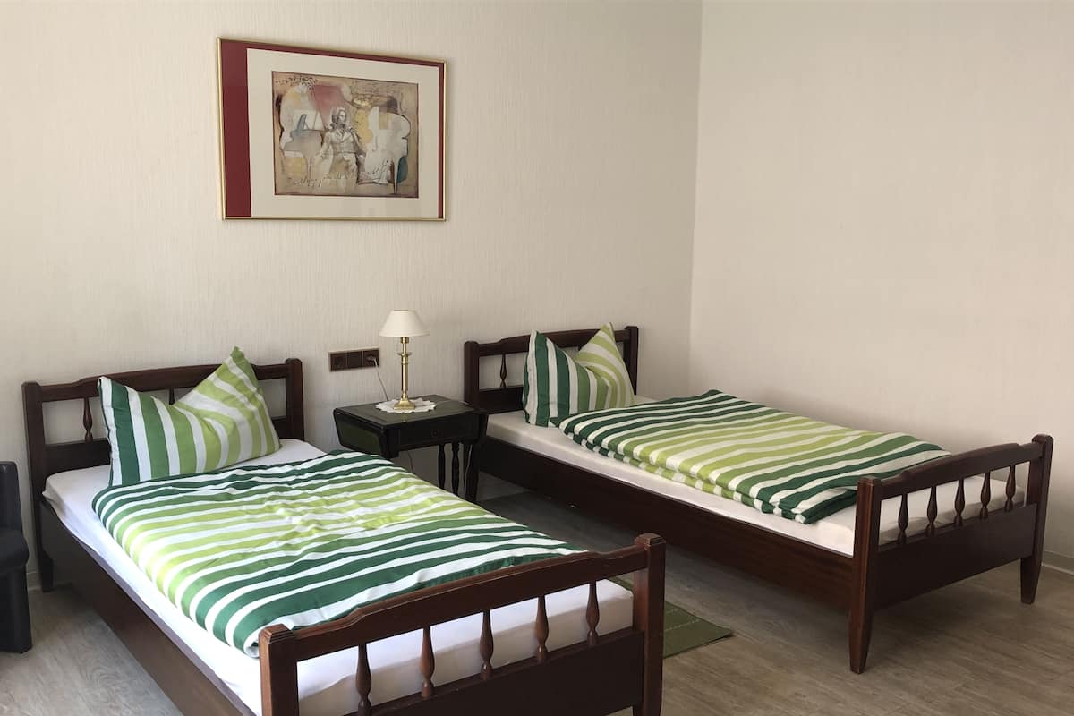 Dos camas individuales con estructura de madera y ropa de cama a rayas verdes, una mesita de noche con lámpara y una obra de arte enmarcada en la pared.