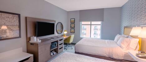 Habitación de hotel con dos camas, televisión, escritorio con lámpara, cómoda y ventana con vistas a edificios.
