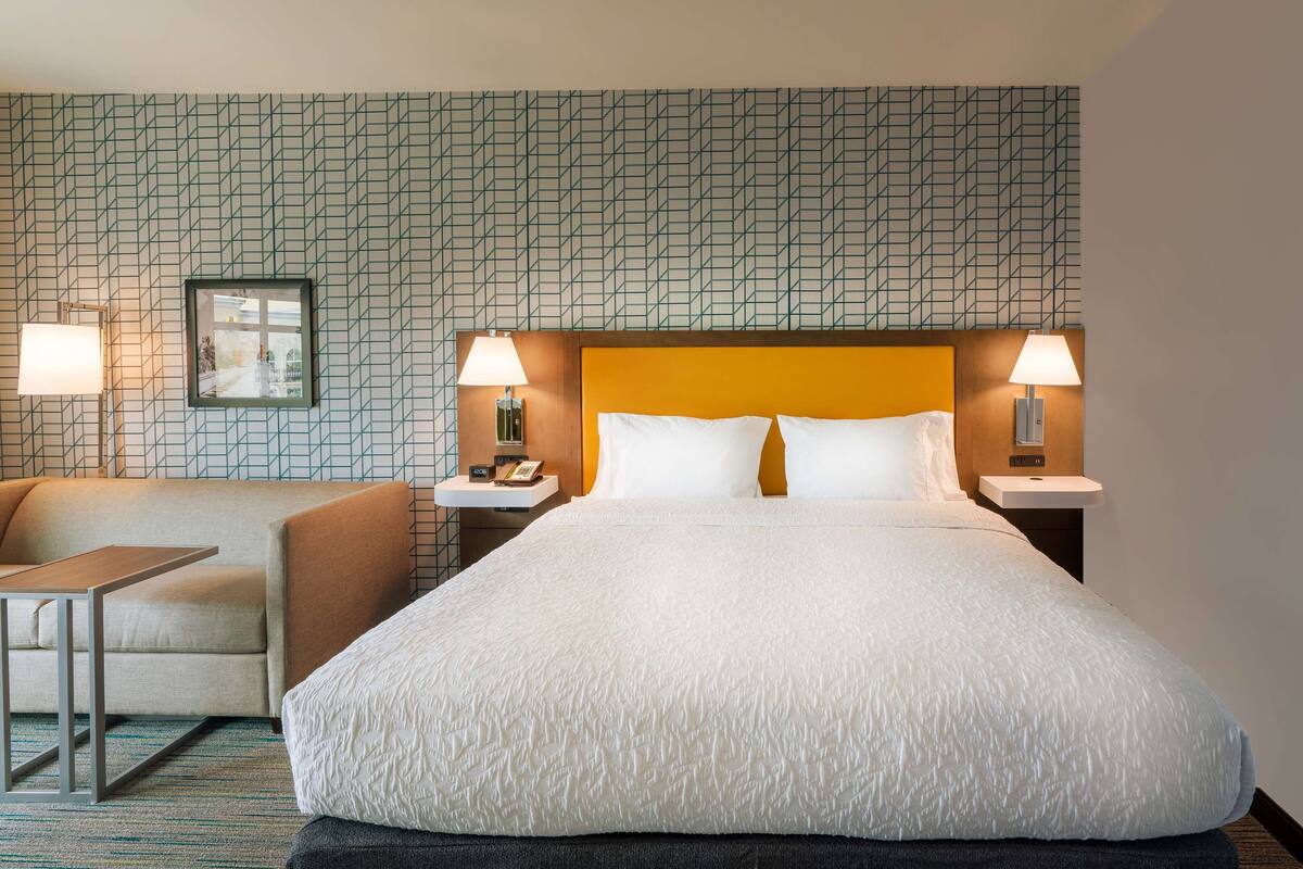 Une chambre d’hôtel avec un grand lit, deux tables de chevet avec des lampes, un petit canapé et un mur décoré d’un motif.