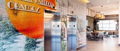 A modern hotel lobby with a large mural, two elevators, and a dining area with striped chairs.