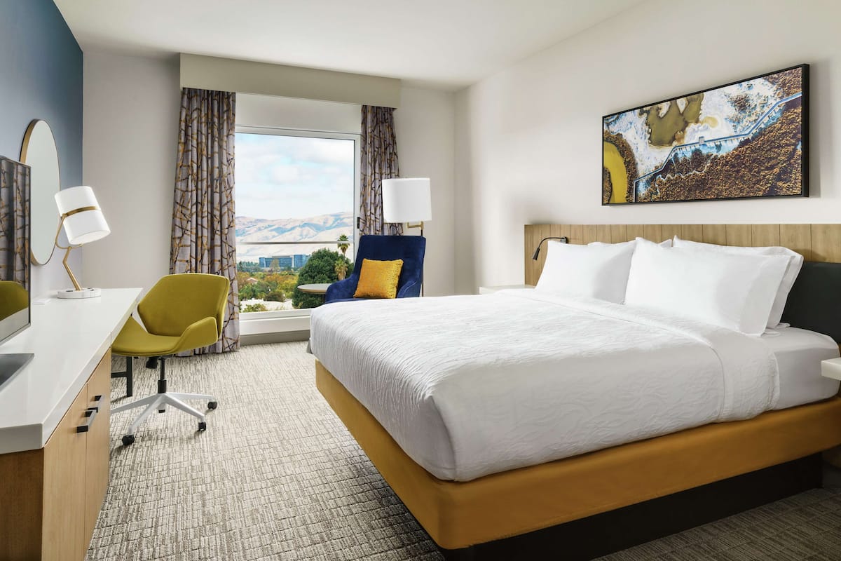 A hotel room with a large bed, a desk with a chair, a window with curtains, and a painting on the wall.