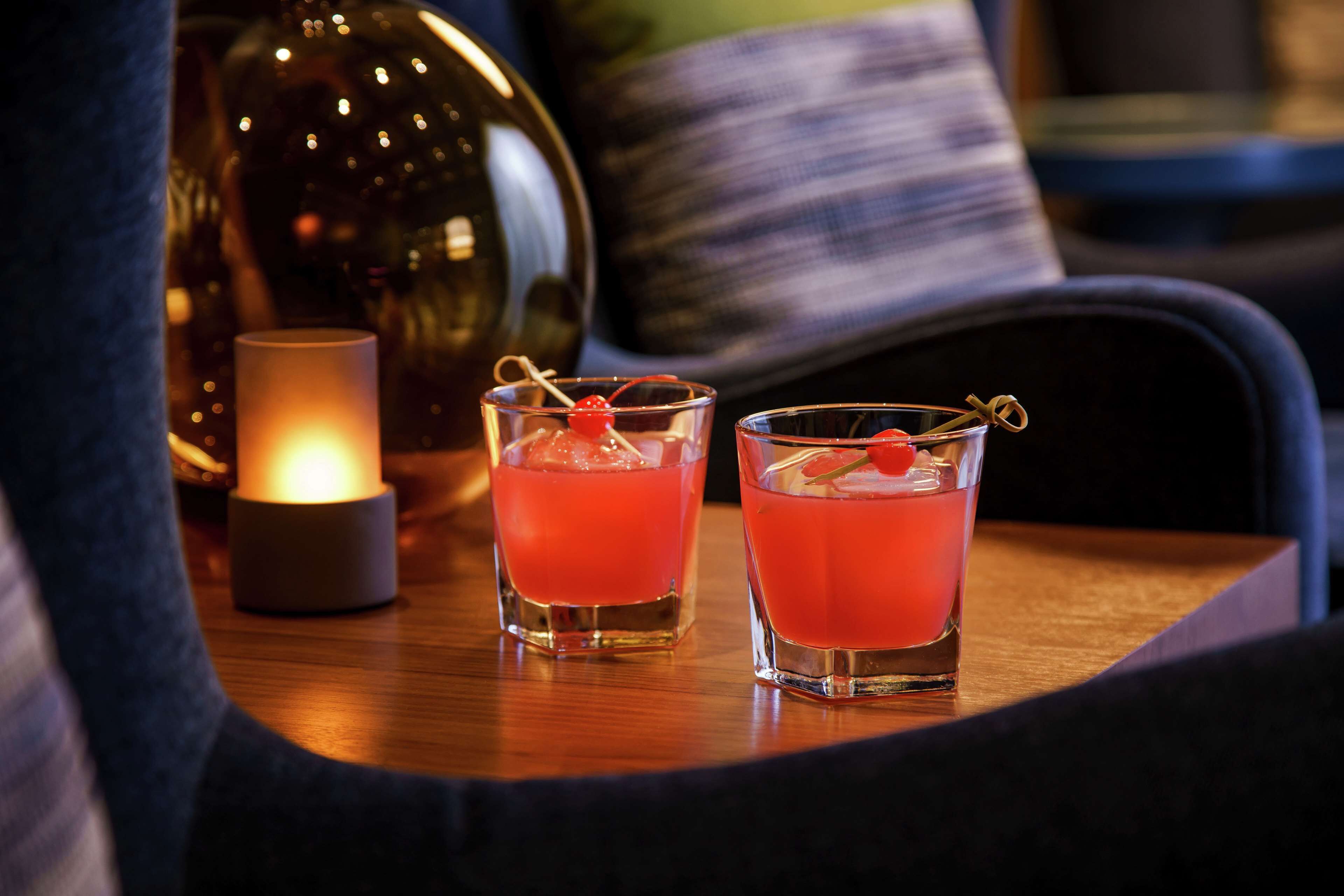 Two glasses of red beverage with cherries on a wooden table, a lit candle, and a dark-colored chair in the background.