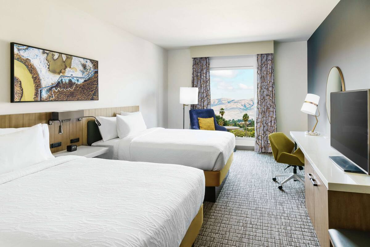 A hotel room with two beds, a desk with a TV, a chair, and a large window with a view.