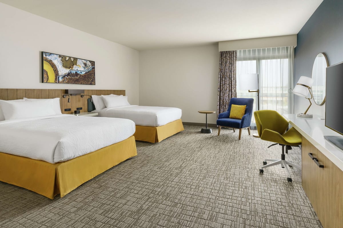 A hotel room with two beds, a desk, a chair, a TV, and a painting on the wall.