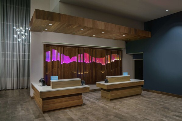 A hotel reception area with two wooden reception desks, a modern light fixture, and a wall with a purple light display.
