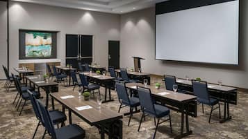 Un salón de conferencias con varias mesas y sillas, una pantalla de proyector y un cuadro grande en la pared.