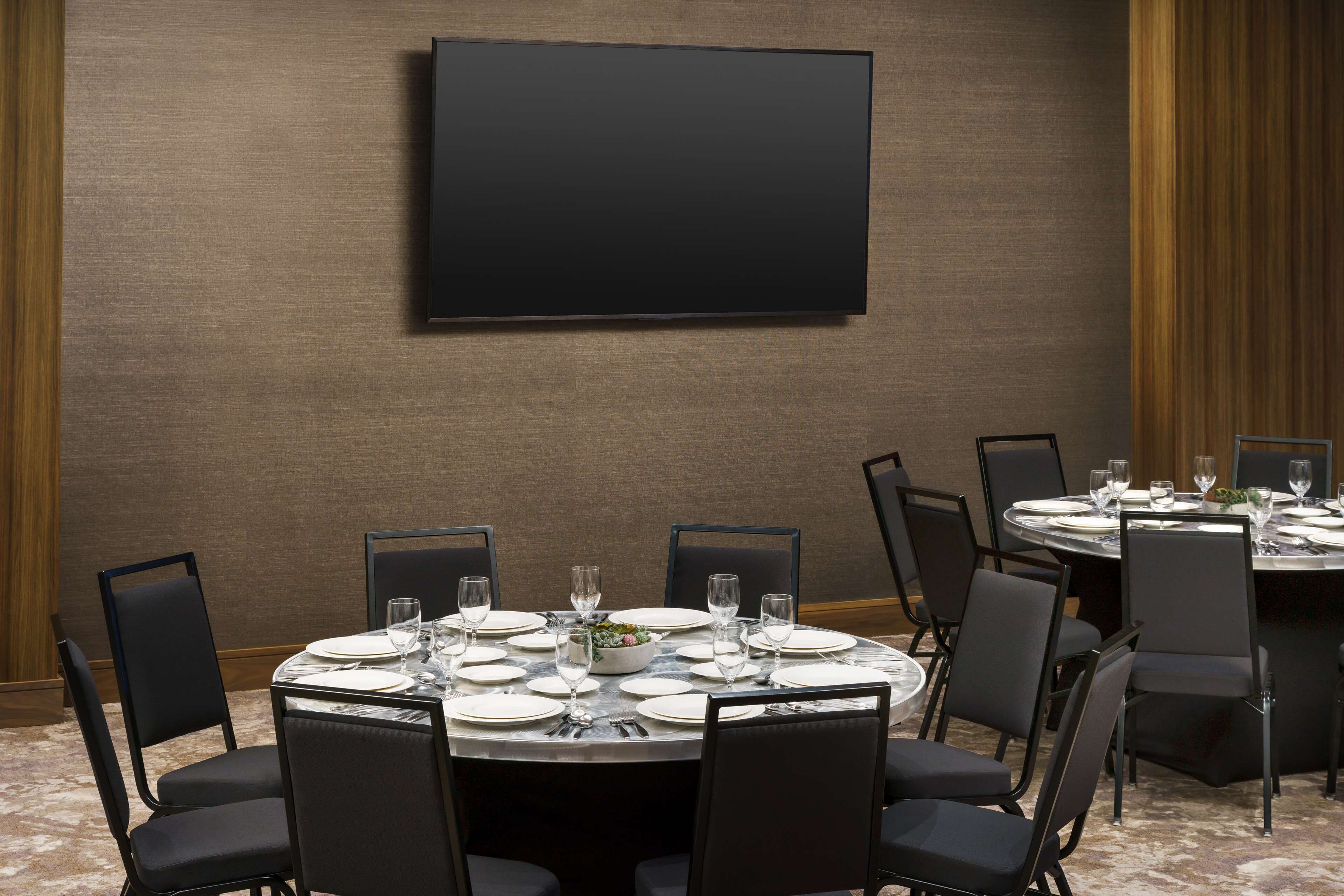 Una sala de conferencias con una gran televisión de pantalla plana montada en la pared, mesas redondas con sillas y una mesa de buffet con platos y vasos.