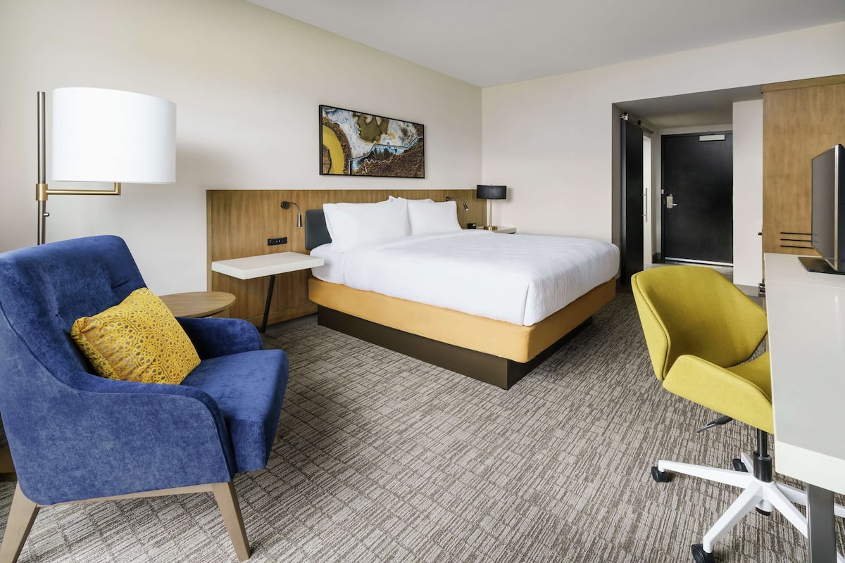 A hotel room with a bed, a blue armchair with a yellow pillow, a yellow office chair, a small table, and a wall-mounted TV.