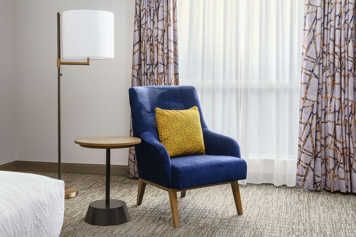 A hotel room with a blue armchair, a small round table, a floor lamp, and patterned curtains.