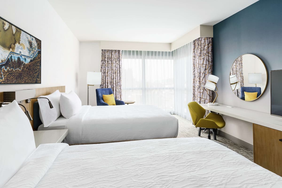 A hotel room with two beds, a desk, a chair, a TV, and a large painting on the wall.
