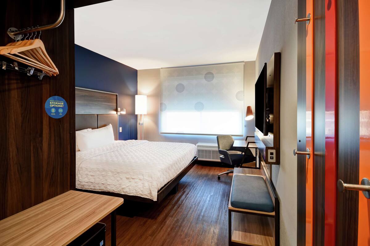 Habitación de hotel con cama, escritorio, silla y televisor. Tiene una ventana con vistas, un televisor montado en la pared y un cartel con información sobre opciones de almacenamiento.