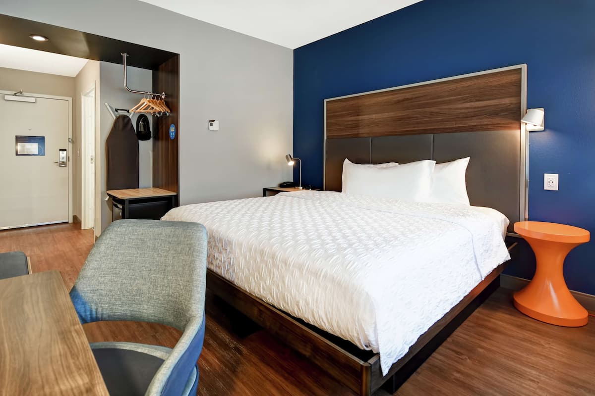 Habitación de hotel con una cama grande, una mesita de noche de madera, una silla, una pequeña mesita auxiliar naranja y un perchero con ganchos.