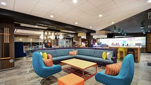 A modern lobby with a sectional sofa, two blue armchairs, a wooden coffee table, and a small orange ottoman. - Tru By Hilton Cincinnati Airport South Florence (Florence)
