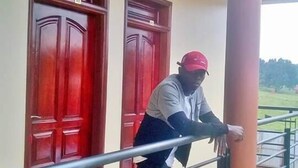 A person leaning on a metal railing outside a building with two red doors and a beige exterior. - Kiboga Resort Hotel (Kiboga)