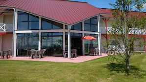 Een gebouw met twee verdiepingen, een rood dak, grote glazen ramen en terras met stoelen en een parasol.