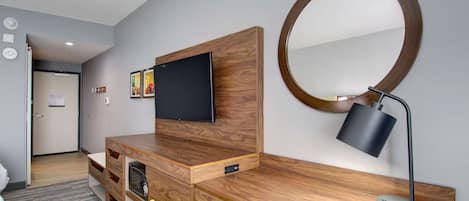 A modern hotel room with a wooden desk, a flat-screen TV, a round mirror, and a desk lamp.