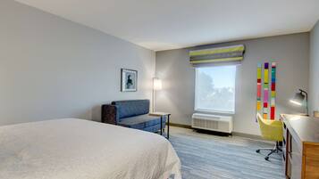 A hotel room with a large bed, a desk with a chair, a window with a view, a wall-mounted air conditioner, and a colorful artwork on the wall.