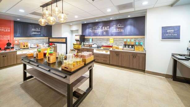 Um café moderno com uma mesa central de atendimento, área de cozinha e parede decorada com temas gastronômicos.