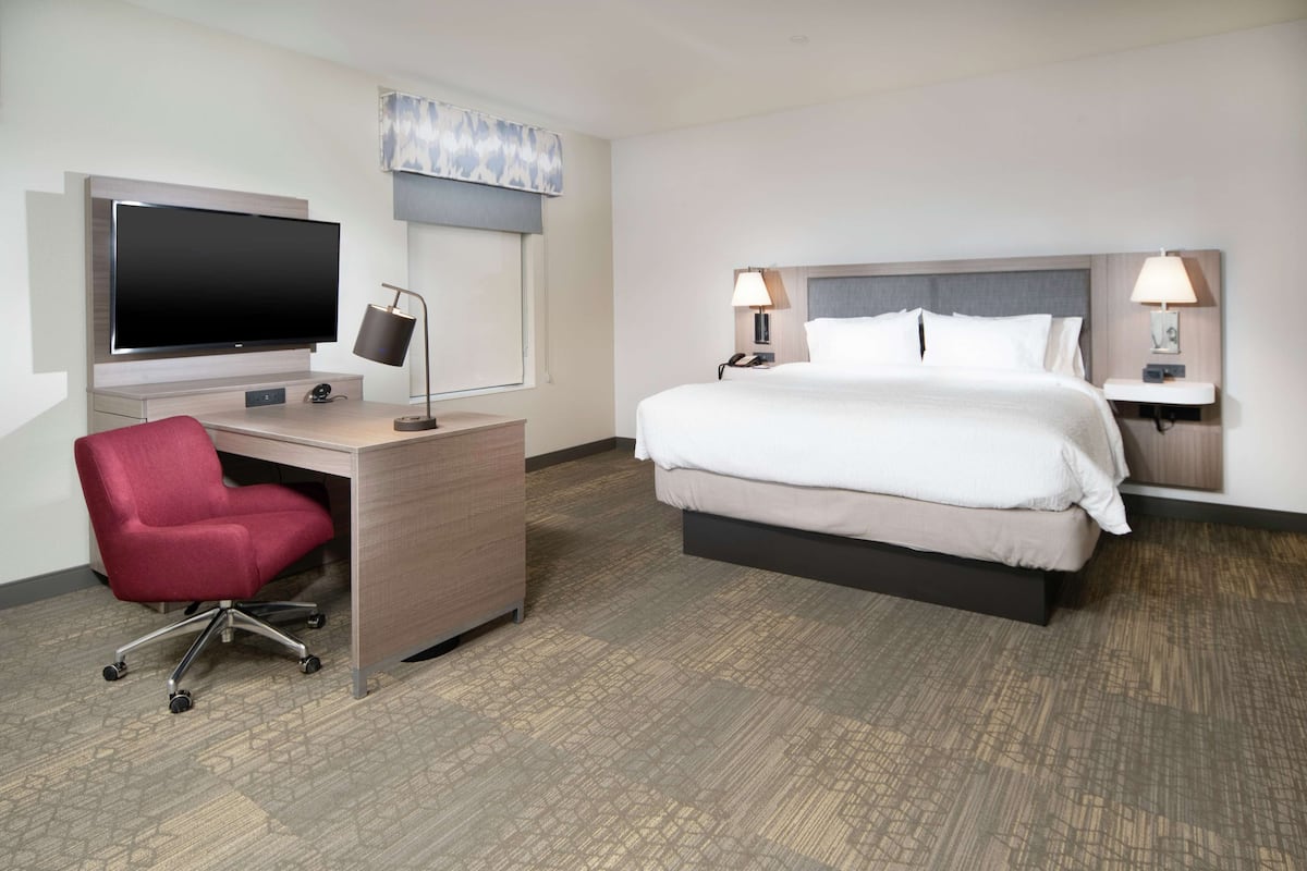 Quarto de hotel com cama, escrivaninha, cadeira, televisão e luminárias.
