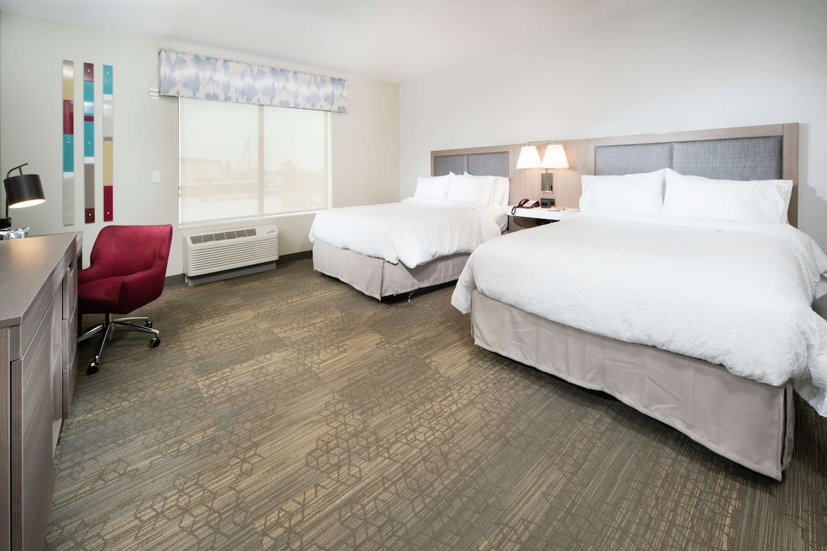 Quarto de hotel com duas camas, uma escrivaninha, uma cadeira, uma janela com vista e um painel decorativo na parede.