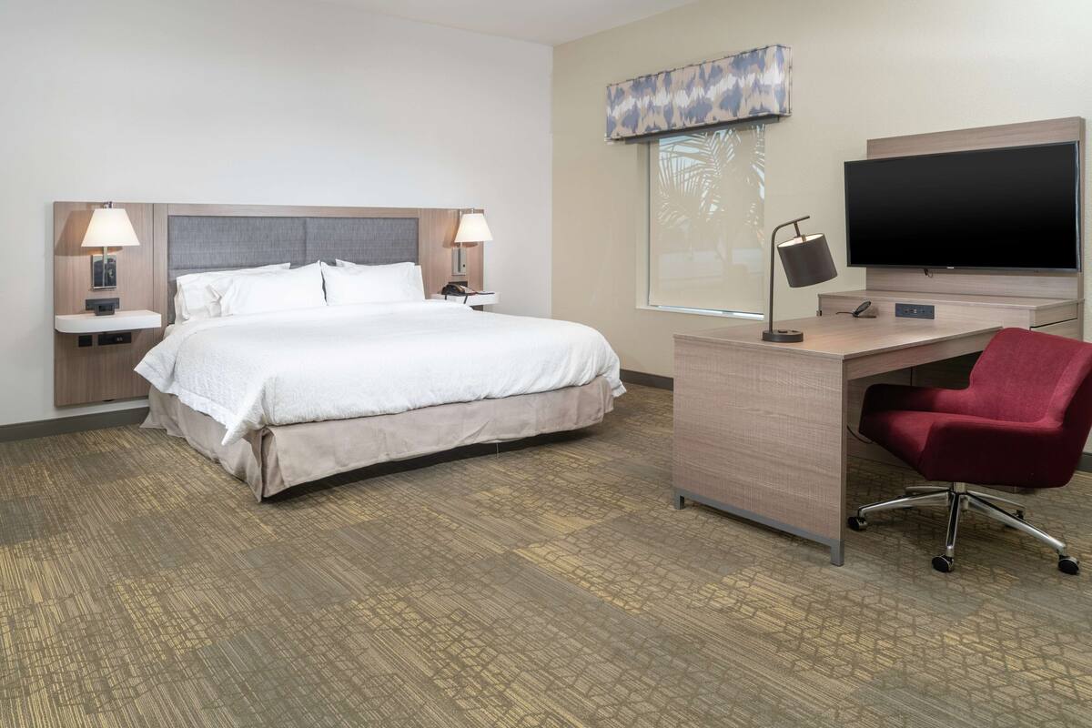 Quarto de hotel com uma cama grande, uma escrivaninha com cadeira, televisão de tela plana e janela com vista para palmeiras.