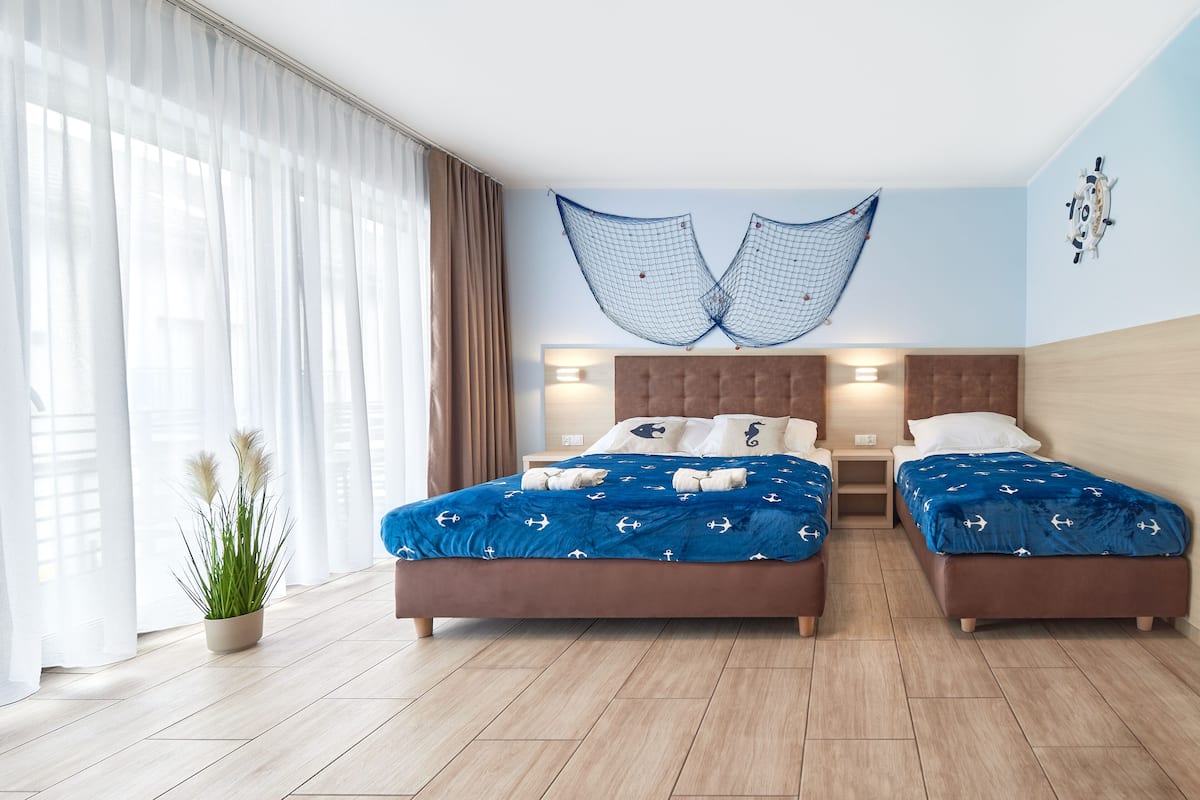 Ein Doppelbett mit blauer Bettdecke, zwei Nachttische, ein Topf mit Pflanze und ein großes Fenster mit Vorhängen.