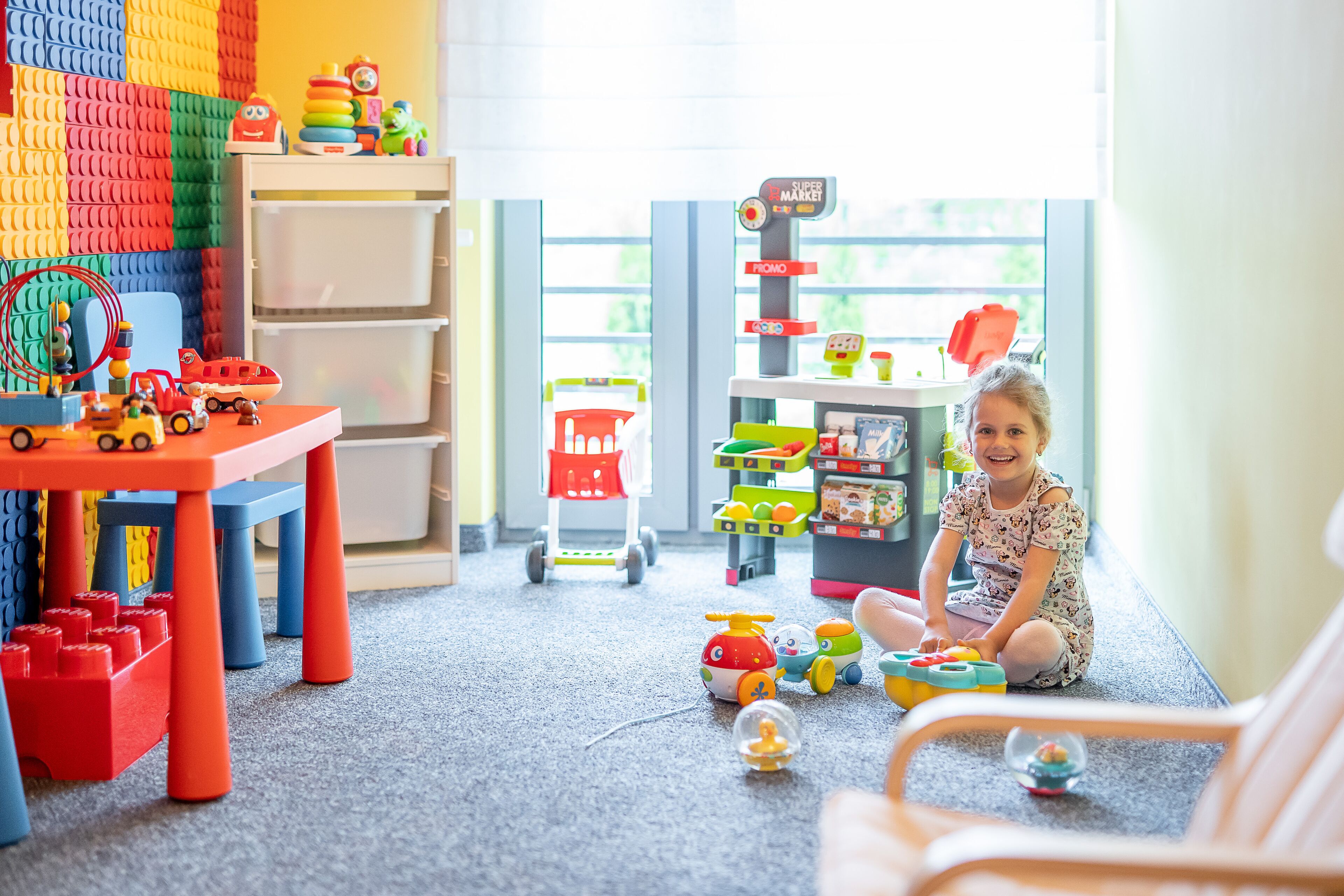 Een kind speelt met speelgoed in een kamer met een kleurrijk speelgedeelte, planken vol speelgoed en een raam met uitzicht.
