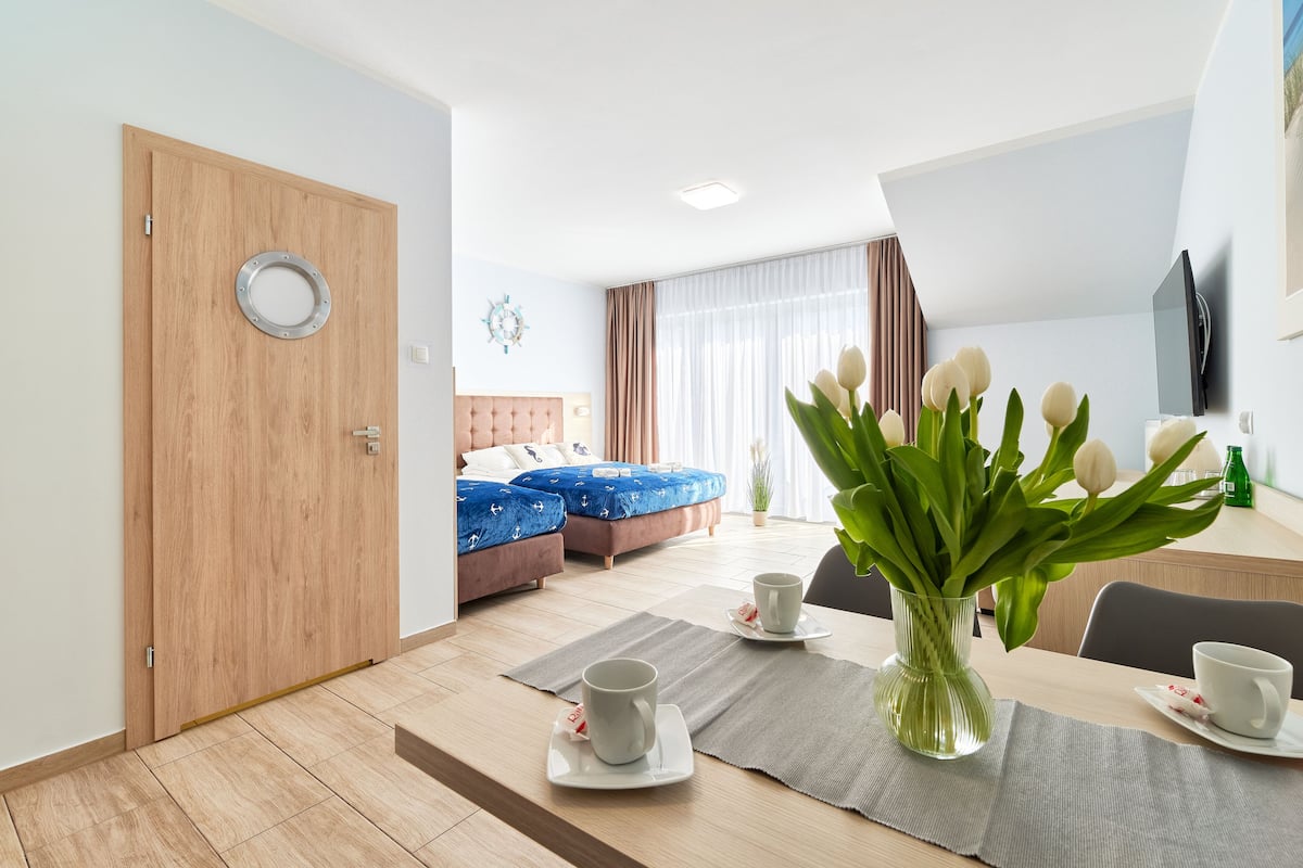 Ein modernes Hotelzimmer mit Holztür, einem Bett mit blauer Bettdecke, einem Essbereich mit Tisch für zwei Personen und einer Vase mit weißen Blumen.