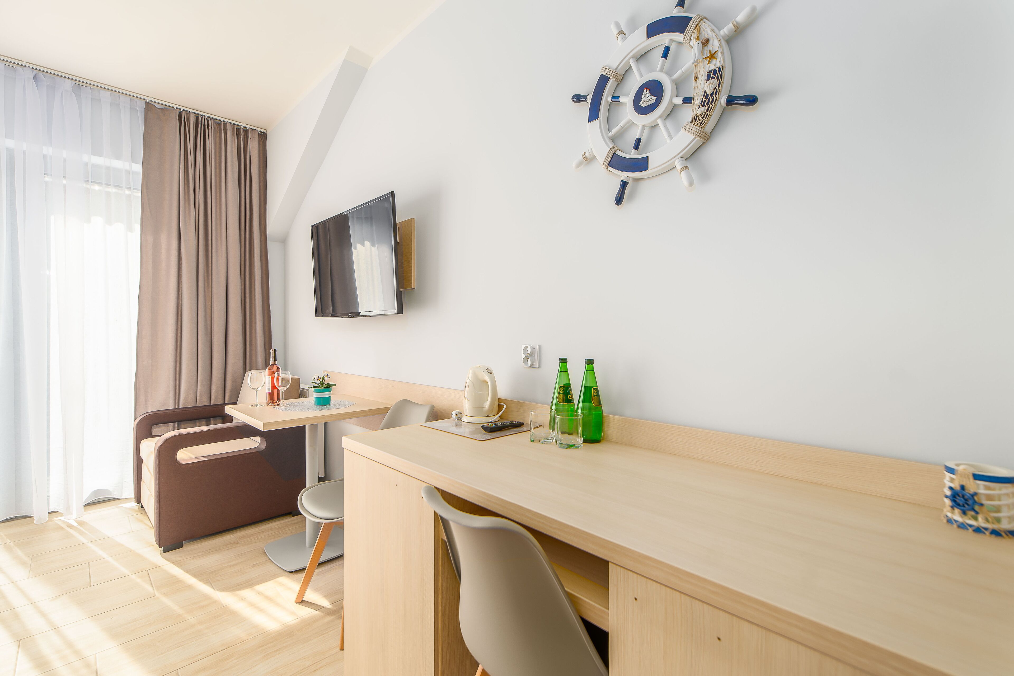 Una habitación de hotel moderna con un escritorio de madera, una silla, una cama, un televisor, una decoración con timón de barco y una ventana con cortinas.