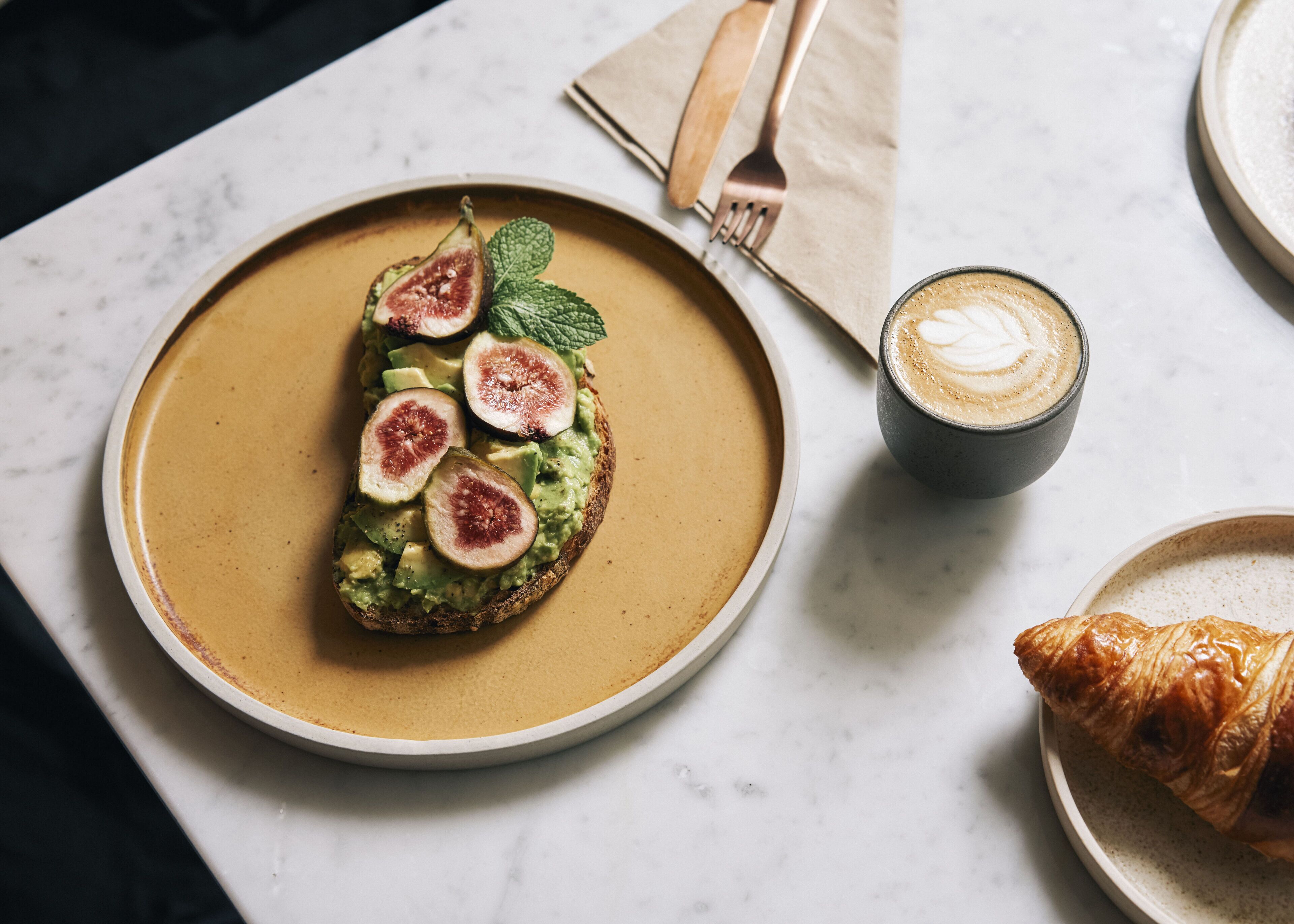 Un plato con tostada de aguacate, higos y hojas de menta, una taza de café y un croissant sobre una mesa de mármol.