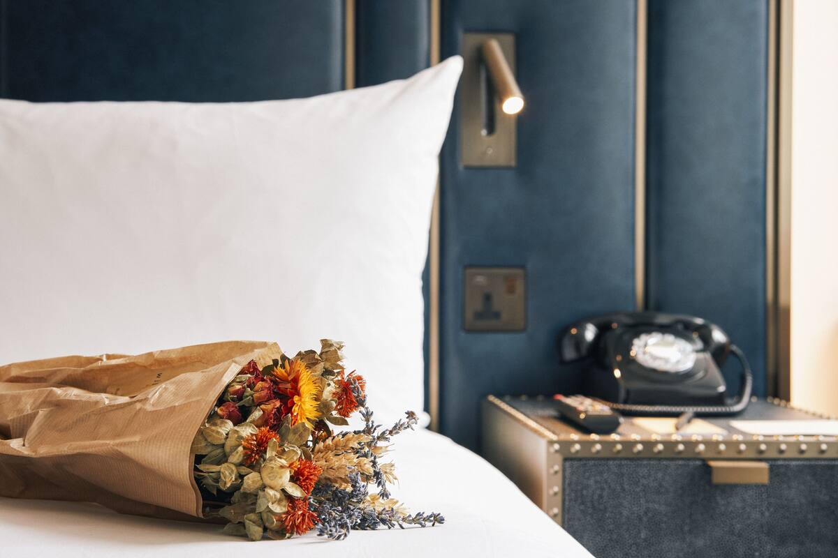 A bed with a white pillow, a bouquet of flowers, a bedside table with a telephone, and a wall with a light switch.