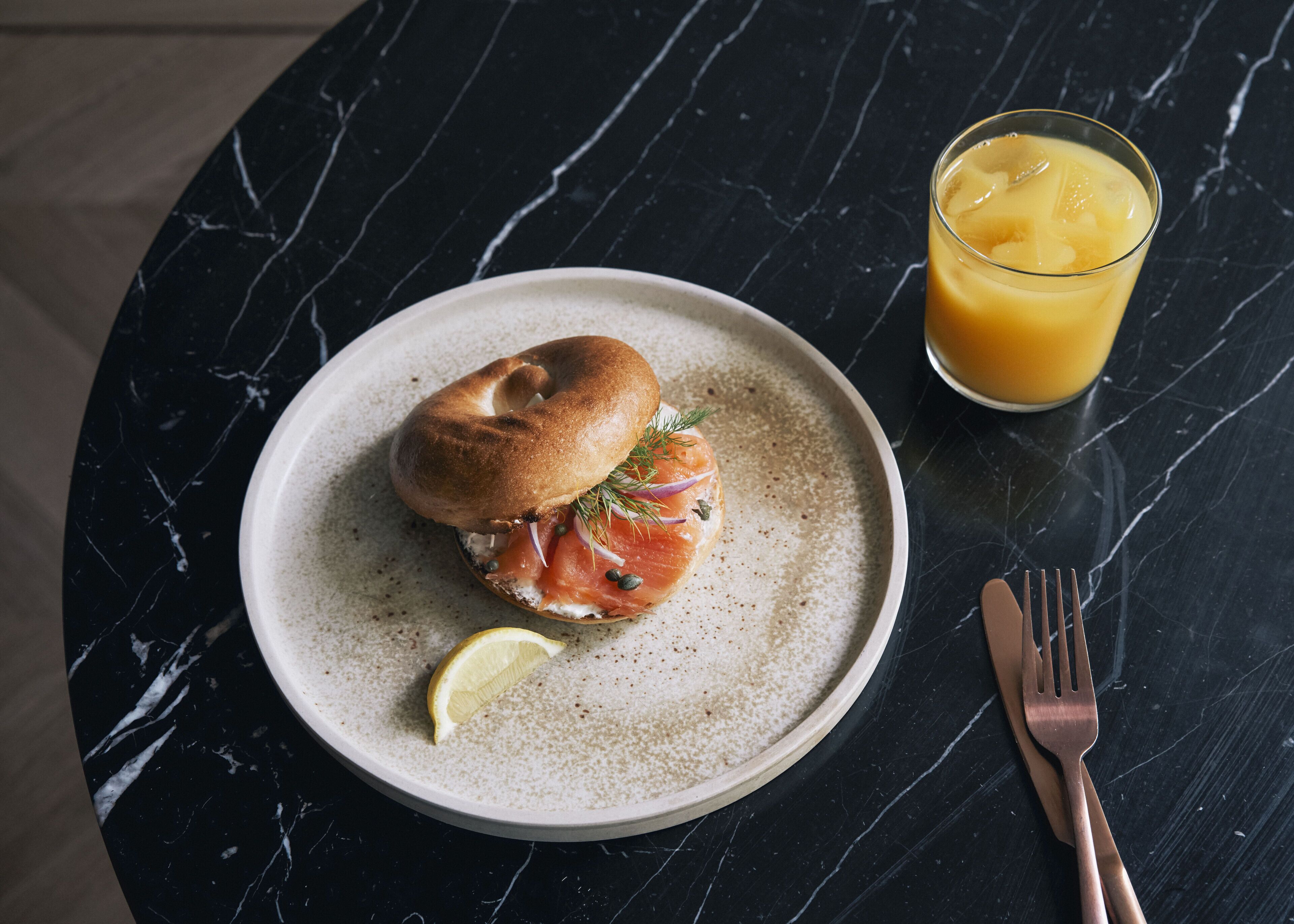 Une assiette contenant un sandwich au bagel, une tranche de citron et un verre de jus d’orange, le tout posé sur une table en marbre foncé.