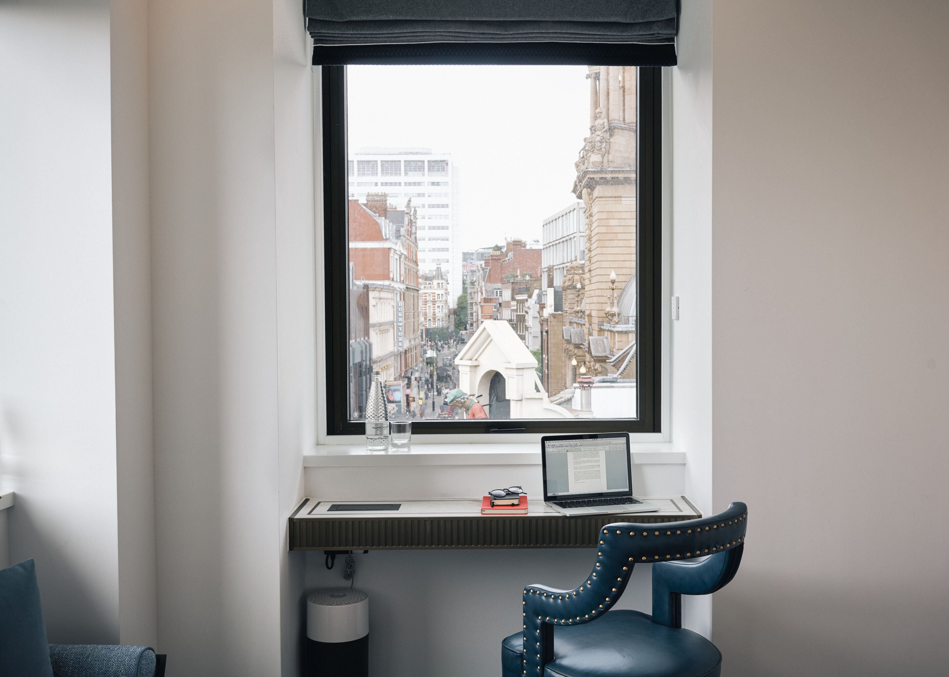 A room with a desk, a chair, a window with a view of a cityscape, and a trash can.