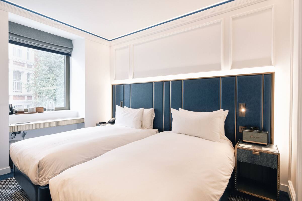 A hotel room with two beds, a large window, a view of buildings, a nightstand with a lamp, and a speaker.