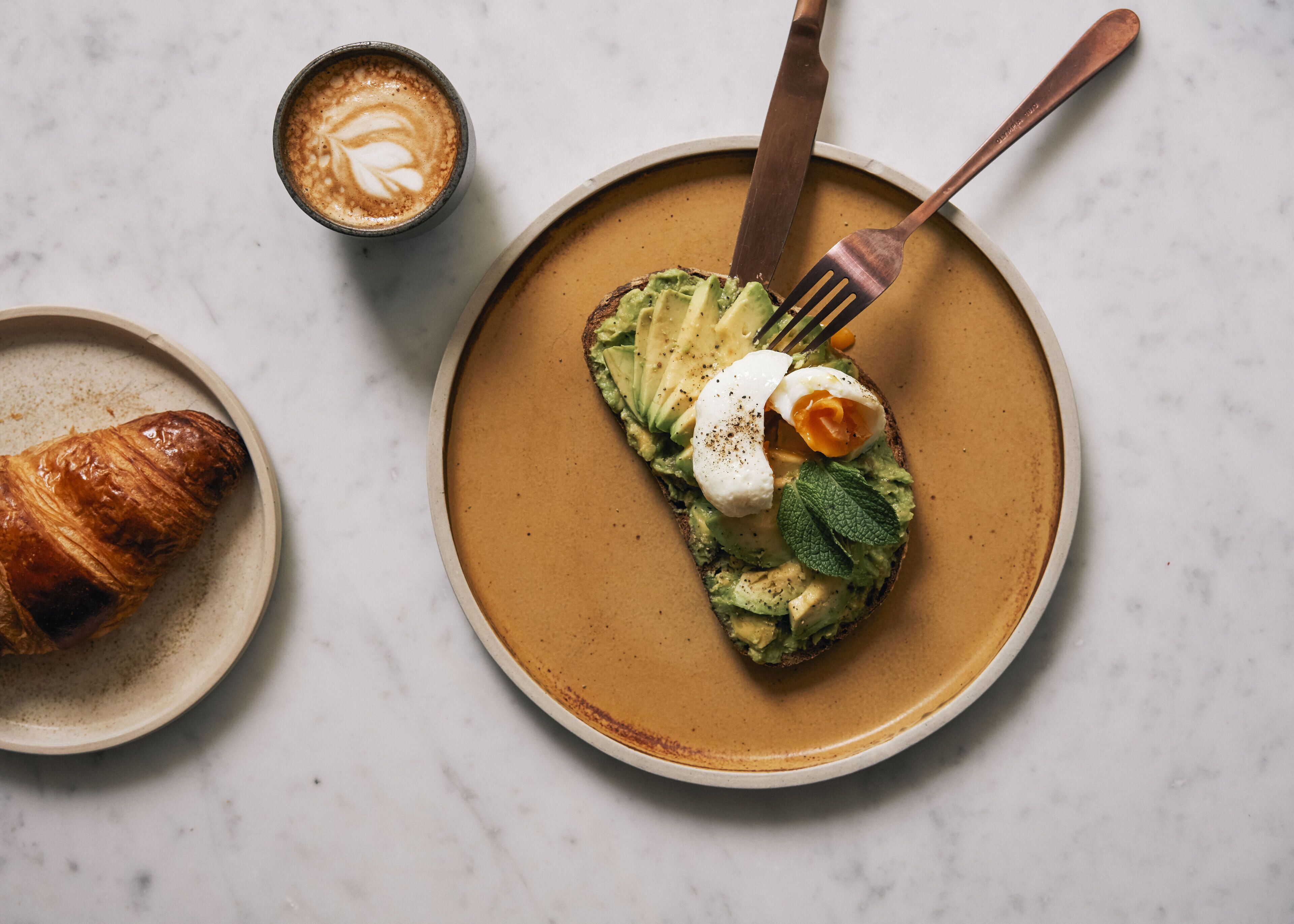 A plate with avocado toast topped with a poached egg and a croissant on the side.