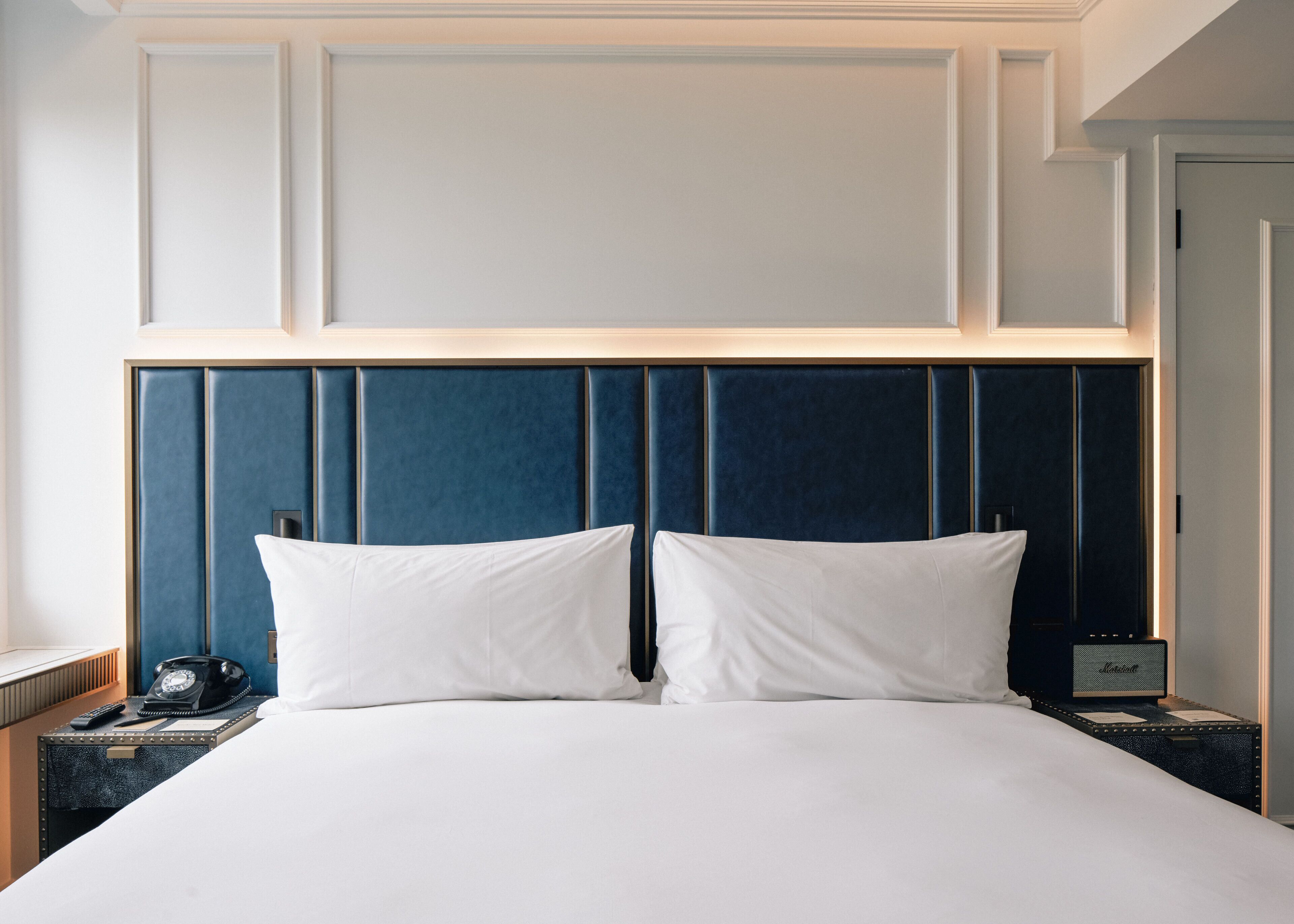 Habitación de hotel con una cama grande, dos almohadas blancas, un cabecero azul oscuro, una mesita de noche con lámpara y un teléfono.