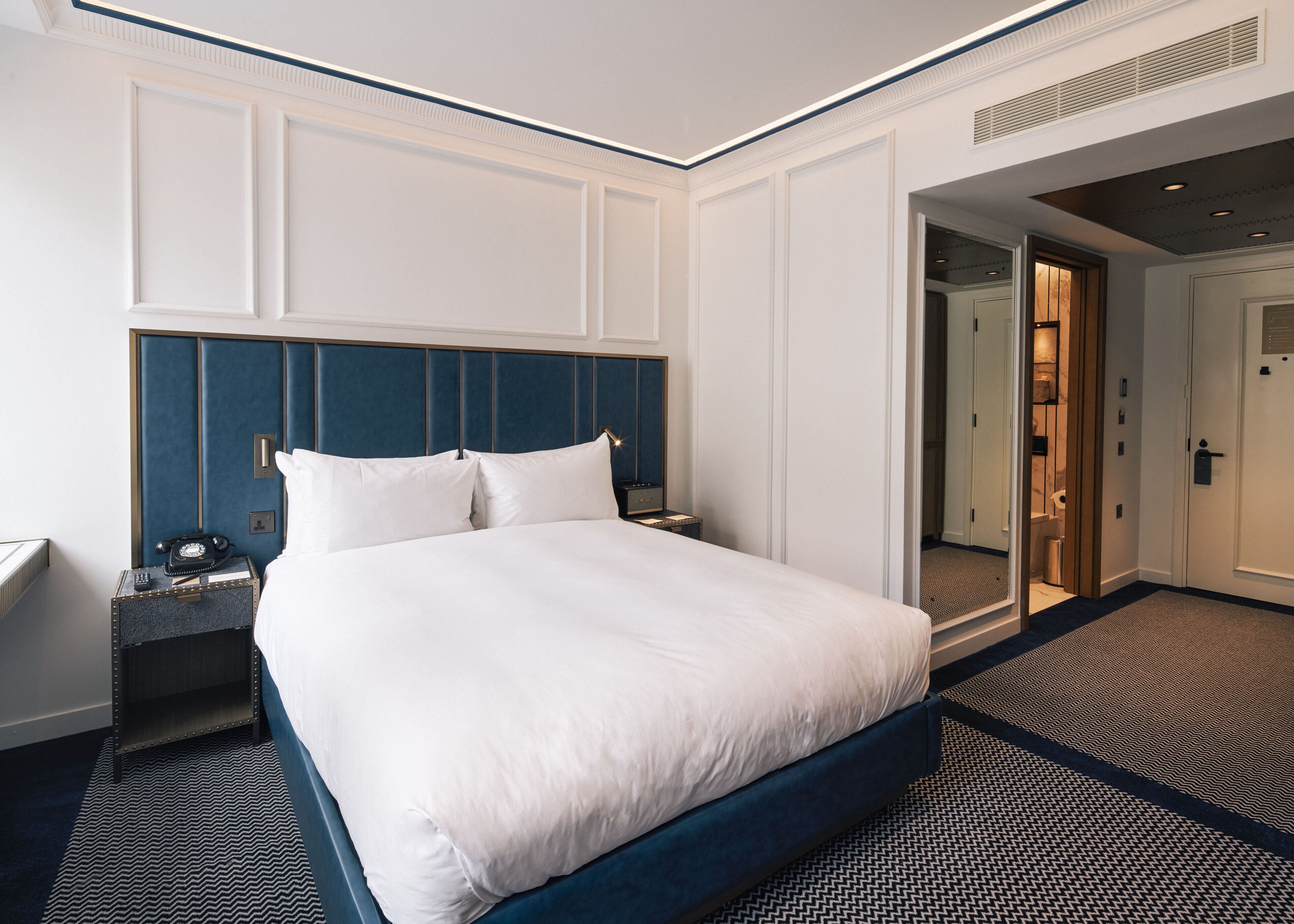 Habitación de hotel con una cama grande, una mesita de noche con teléfono, un espejo y una puerta que da a un baño.