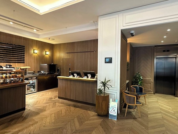 Zona de recepción de hotel con un mostrador de madera, una máquina de café y un cartel que indica suelo mojado.
