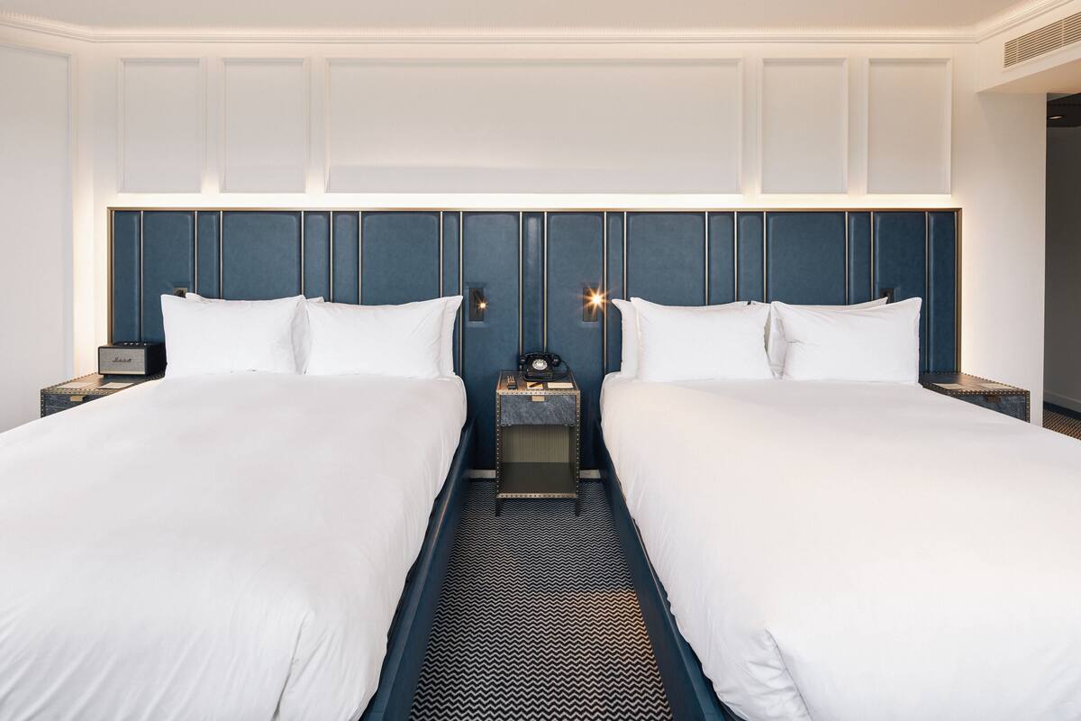A hotel room with two beds, a headboard with vertical lines, a nightstand with a clock, and a small table with a lamp.